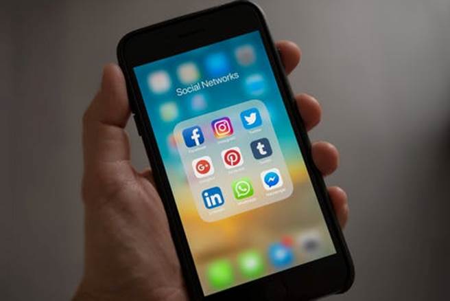 Hand holding a smartphone displaying social media apps including Facebook, Instagram, Twitter, LinkedIn, and WhatsApp on the screen